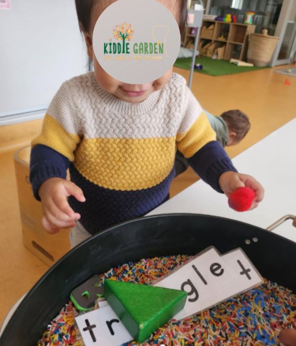 infant at kiddie garden holding a soft pom pom and reaching into coloured rice