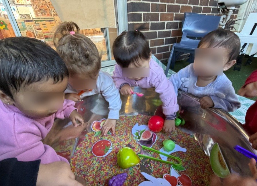 four infants reaching into a sensory bowl with coloured rice and toys, supervised by educators at Kiddie Garden